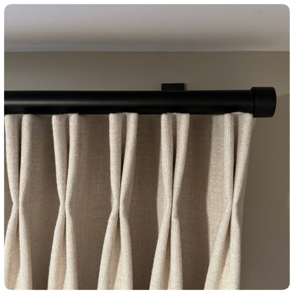 Beige curtains hanging from a black curtain rod against a neutral wall. black designer drapery hardware